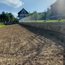 Firma Uslugowa Wojciech Łukaszka - Przygotowany teren pod budowę z widocznymi śladami opon, ogrodzony siatką i betonowym murem. W tle dom z balkonami i zielone drzewa pod błękitnym niebem.