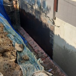 Firma Uslugowa Wojciech Łukaszka - Fundament budynku po izolacji bitumicznej, zabezpieczony folią i drenażem. Widoczny wykop i fragment elewacji z rurą spustową.