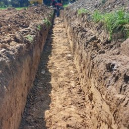 Firma Uslugowa Wojciech Łukaszka - Długi wykop w ziemi, w tle koparka i pracownicy. Wykop świeży, z wyraźnymi śladami pracy maszyny. Praca w toku, teren zielony w tle.