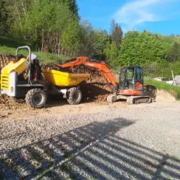 Pomarańczowa minikoparka Kubota U55-4 załadowuje ziemię do żółtej wywrotki Wacker Neuson na placu budowy z utwardzoną żwirową drogą i zielonym tłem drzew.