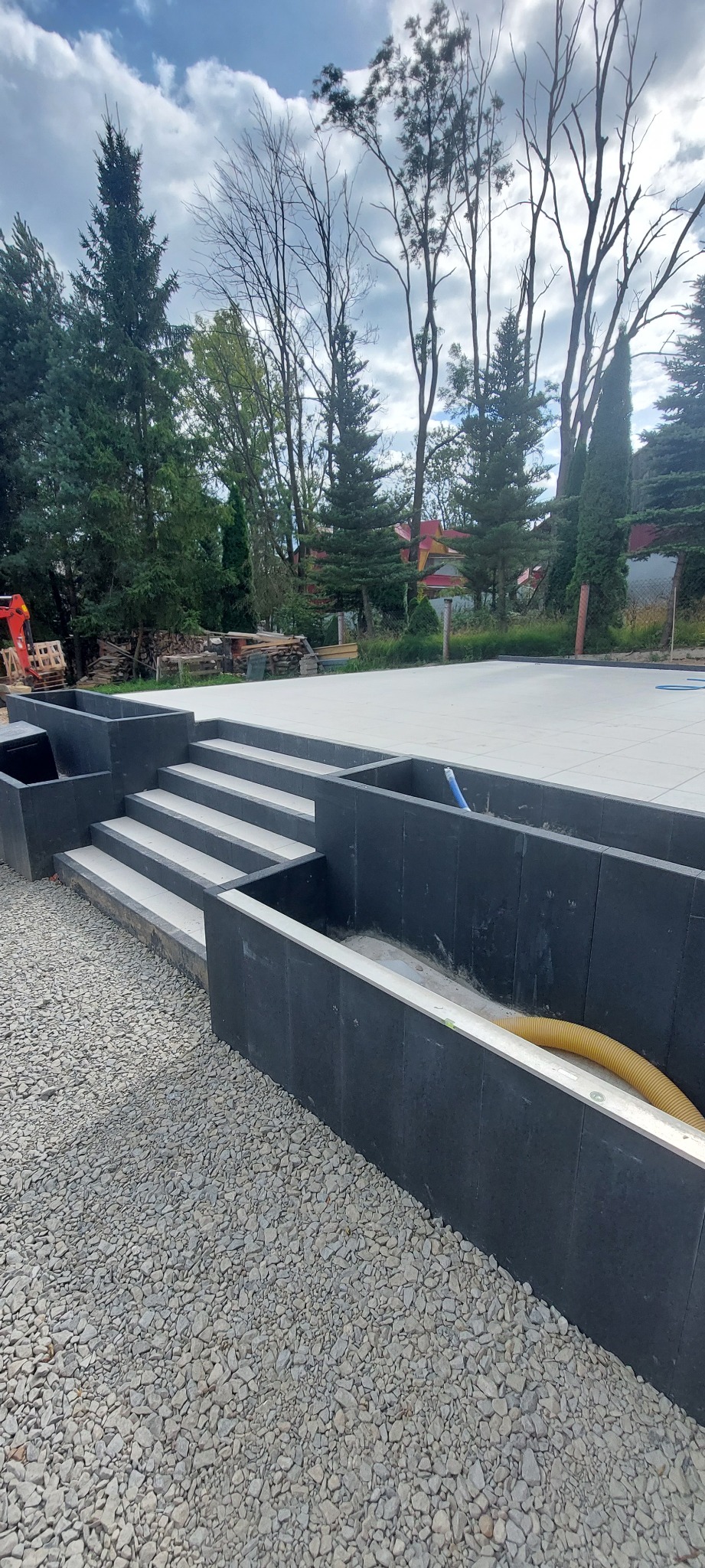 Nowoczesne schody z szarej kostki prowadzące do tarasu, otoczone żwirem i czarnymi ściankami. W tle drzewa i budynek.