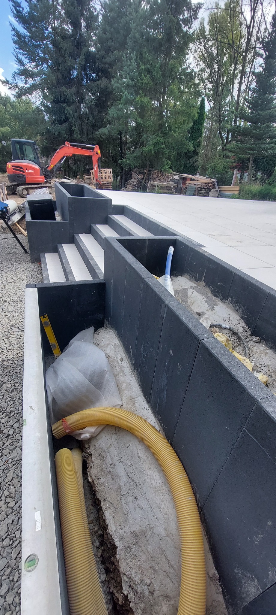 Budowa schodów i murków z bloczków betonowych w Raba Wyżna. W tle widoczny pomarańczowy minikoparka. W wykopie żółte rury drenażowe i poziomica.