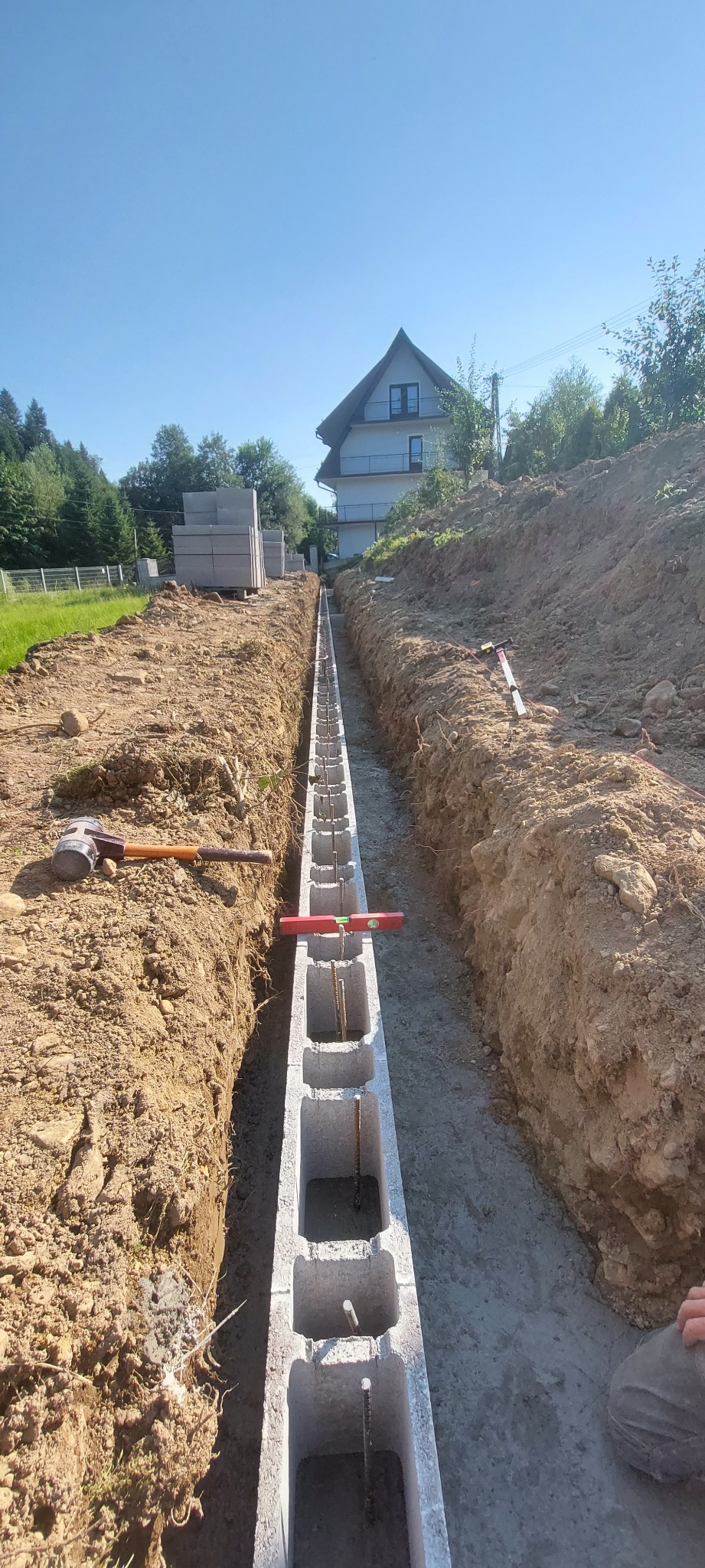 Wykop z betonową wylewką i bloczkami fundamentowymi, zbrojenie pionowe, poziomica, młotek ciesielski w tle, dom jednorodzinny w oddali, słoneczny dzień.