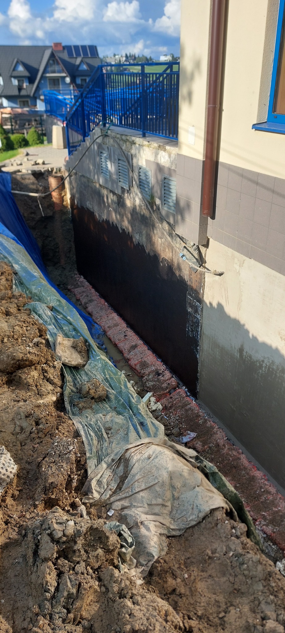 Fundament budynku po izolacji bitumicznej, zabezpieczony folią i drenażem. Widoczny wykop i fragment elewacji z rurą spustową.