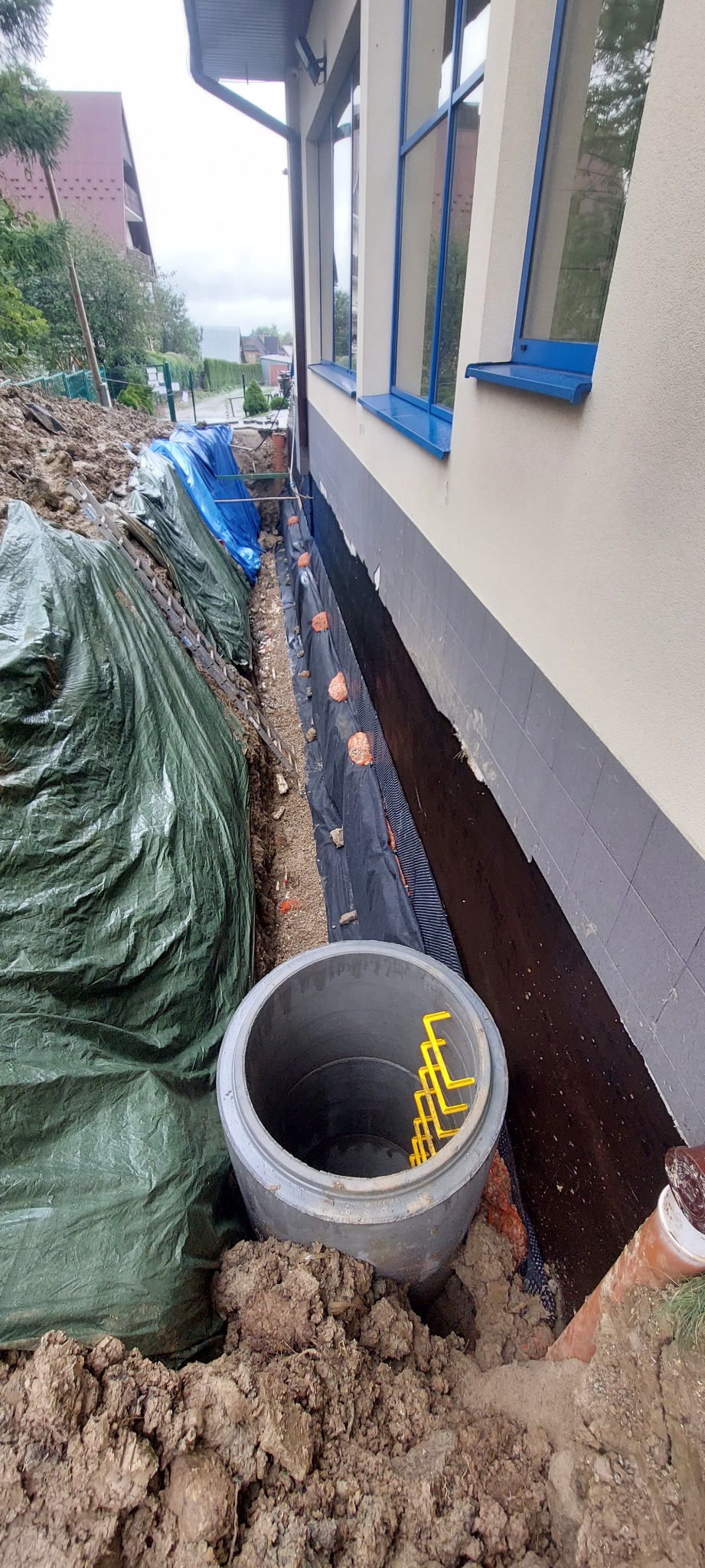 Studzienka kanalizacyjna z żółtymi elementami w wykopie przy budynku, zabezpieczona folią. Widoczna izolacja fundamentów i rury. Prace ziemne w toku.