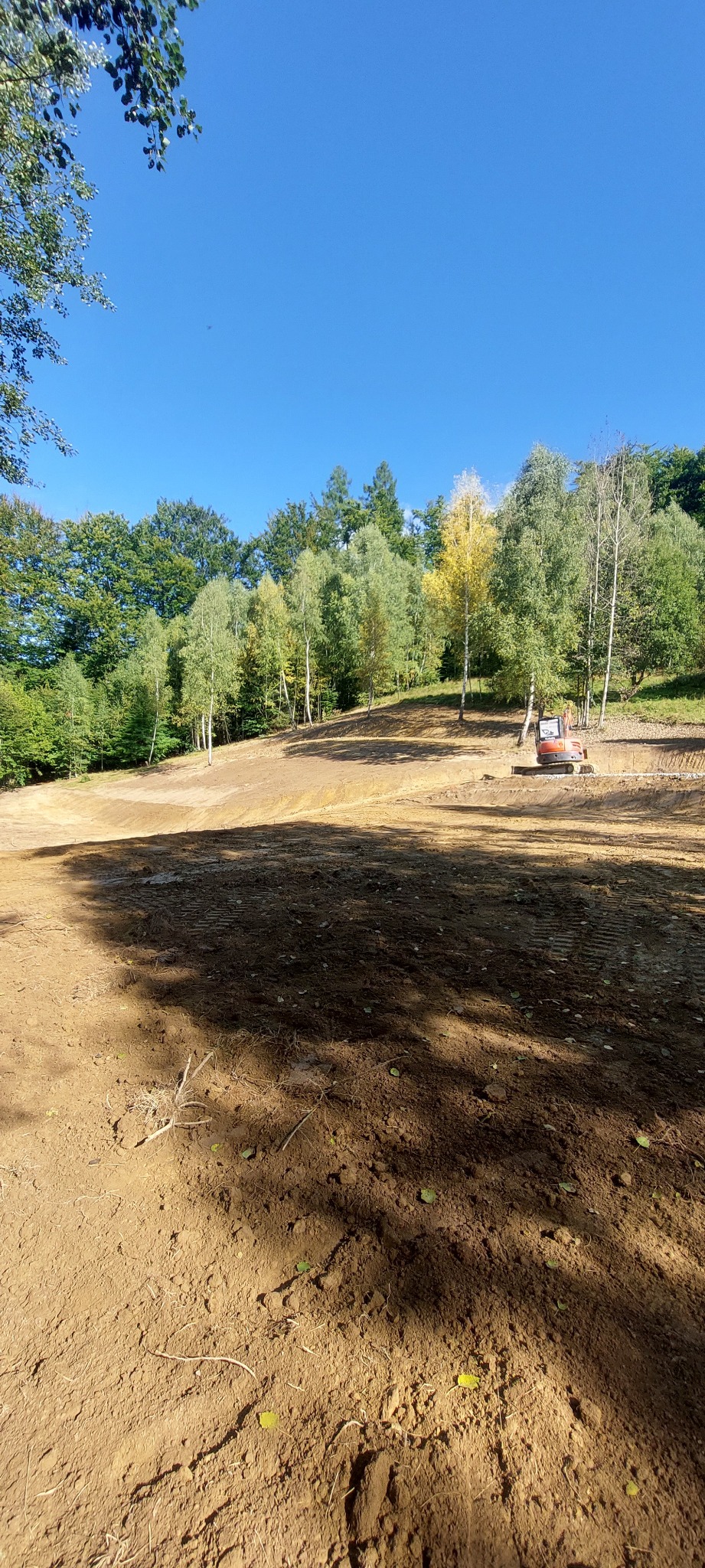 Niwelacja terenu z użyciem koparki na tle lasu i błękitnego nieba. Ślady opon na ziemi wskazują na postęp prac.