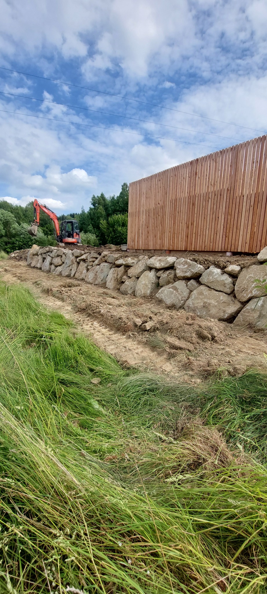 Pomarańczowa koparka przy drewnianej konstrukcji i kamiennym murku oporowym na tle błękitnego nieba z chmurami. Prace ziemne w toku.