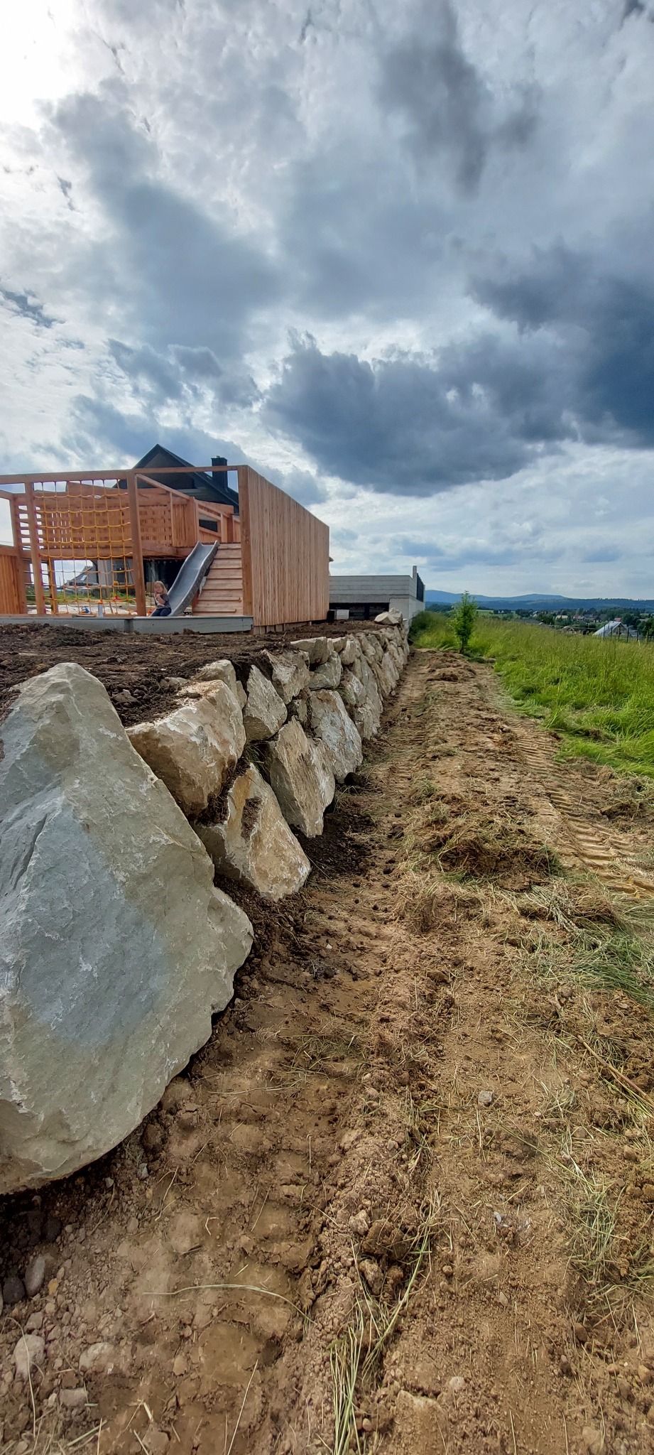 Kamienny mur oporowy na nierównym terenie, z drewnianym placem zabaw w tle i pochmurnym niebem. Widoczne ślady opon na ziemi.
