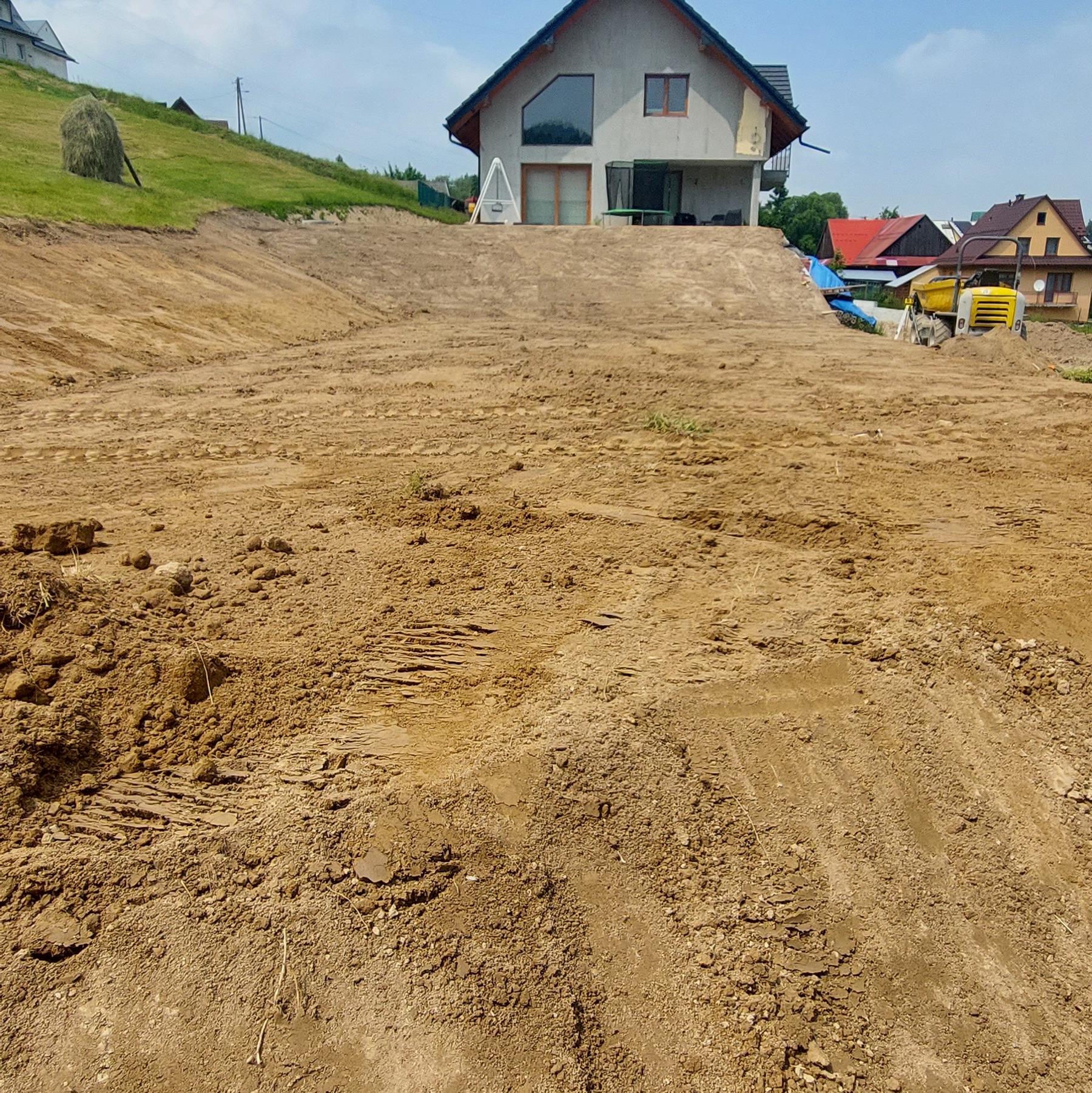 Nierówny teren po niwelacji, z widocznymi śladami opon i budynkiem w tle. Żółty walec drogowy w oddali. Słoneczny dzień.