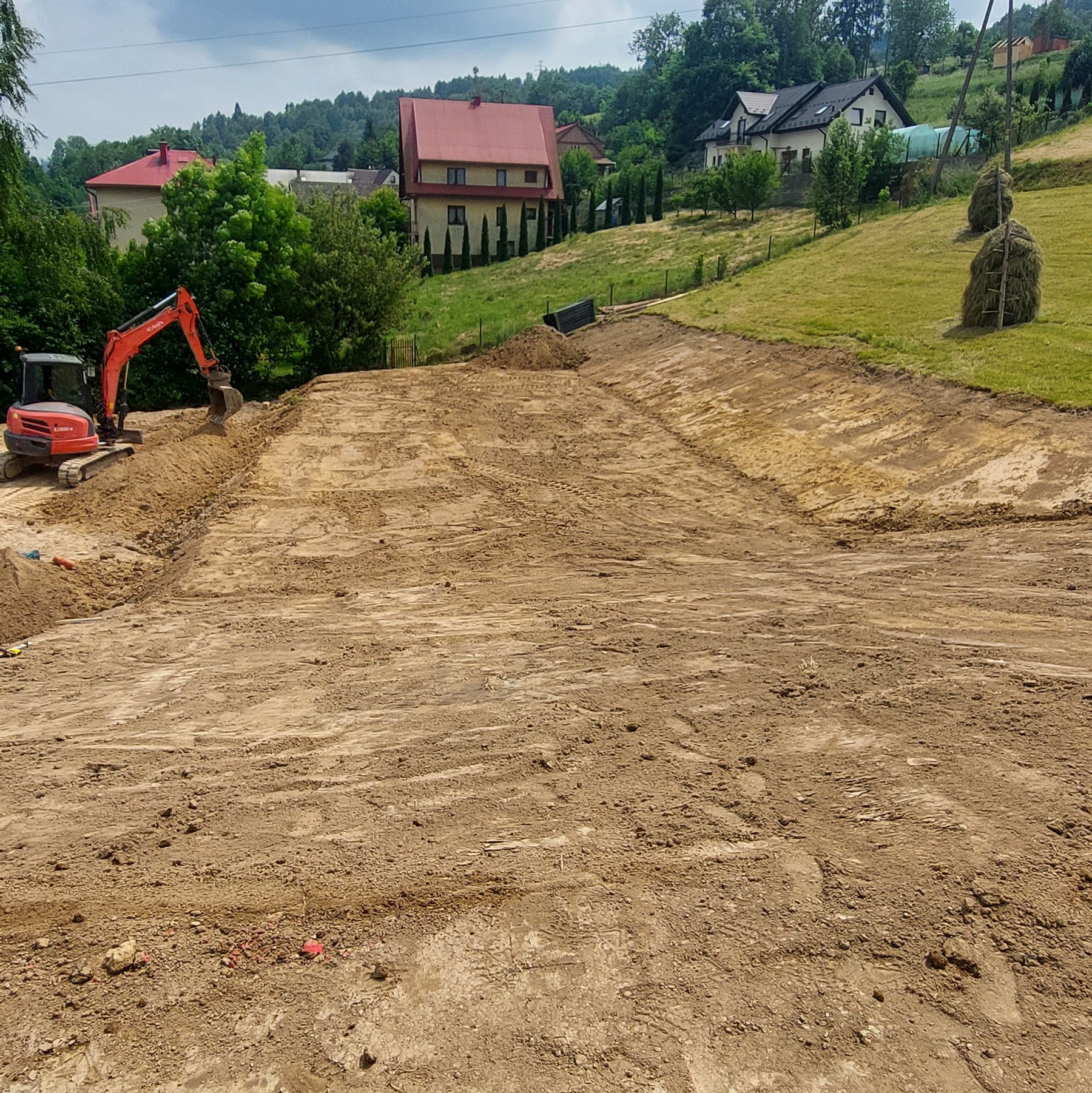 Czerwona koparka Kubota na placu budowy, wyrównany teren pod budowę z widokiem na domy i drzewa w tle, słoneczny dzień.