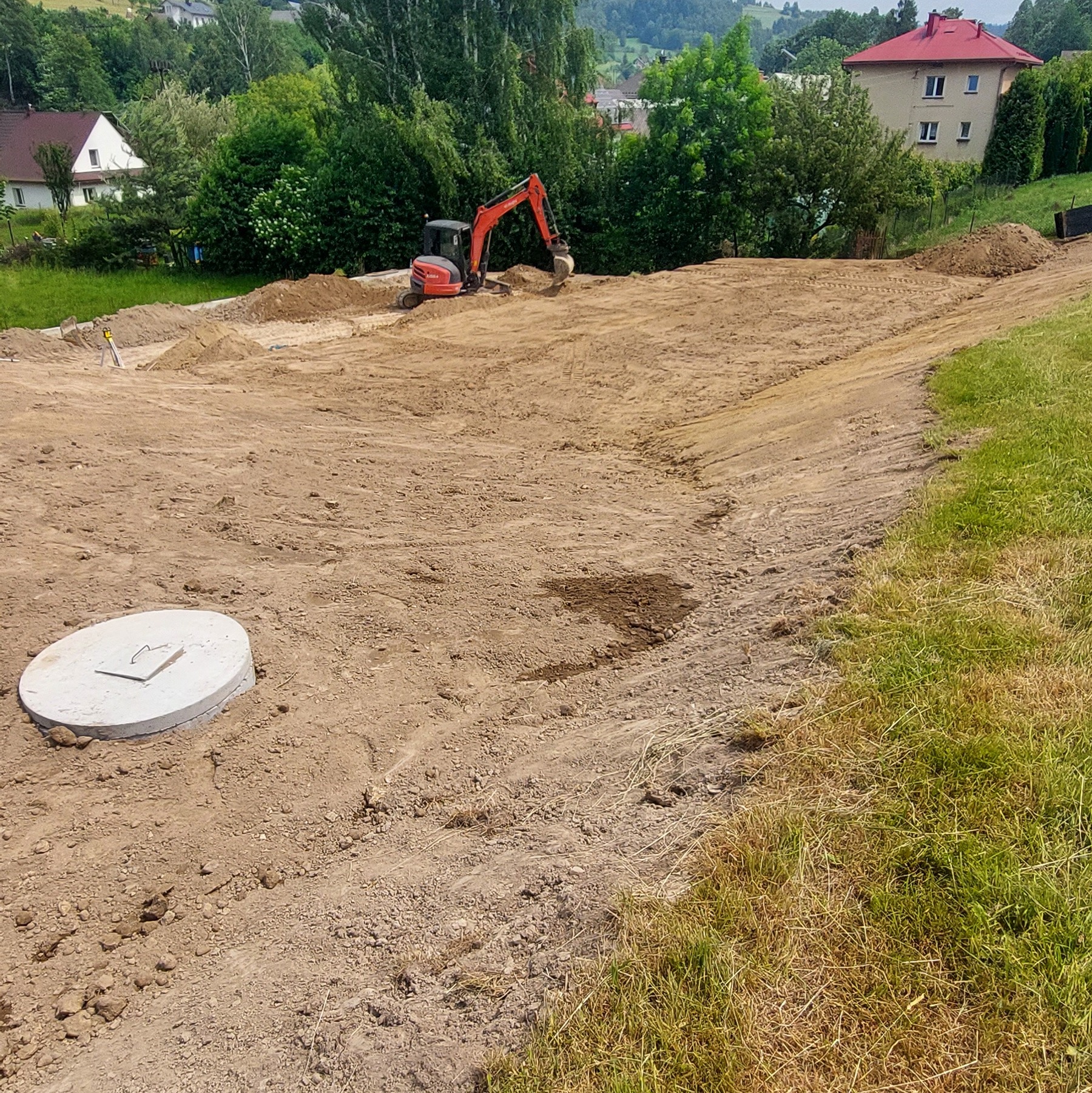 Niwelacja terenu pod budowę z widocznym wykopem, studzienką kanalizacyjną i koparką w tle, otoczoną zielenią i zabudowaniami.