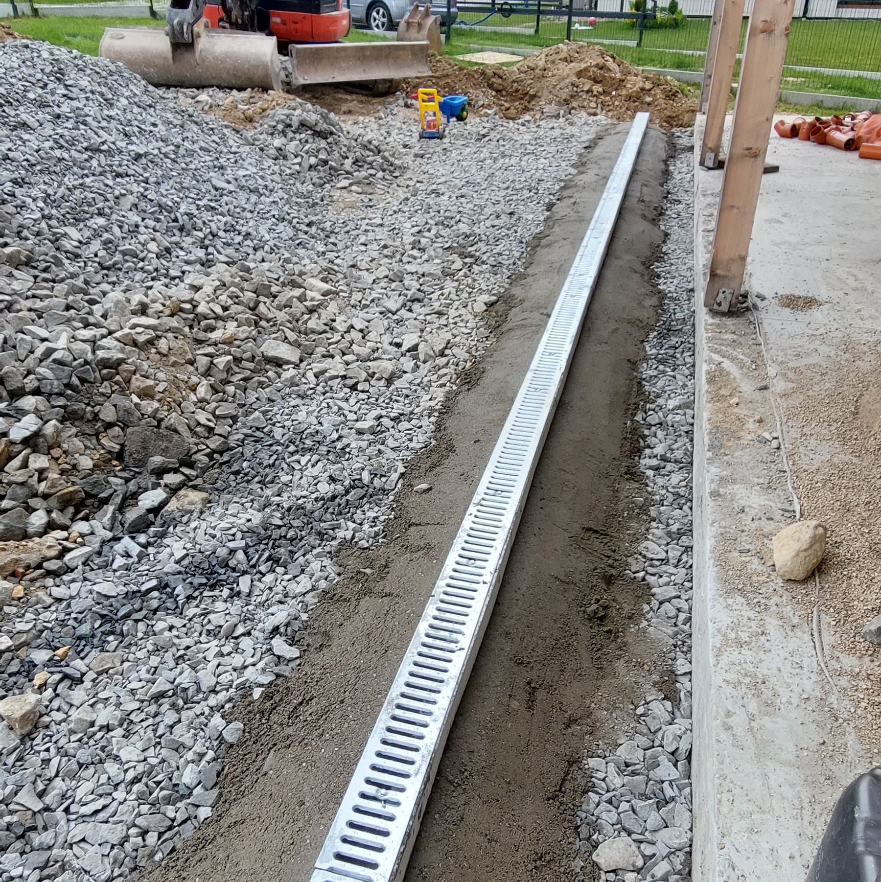 Długi, metalowy odpływ liniowy ułożony w gruncie z kamieniami, obok betonowej konstrukcji. W tle koparka i zabawki dziecięce.