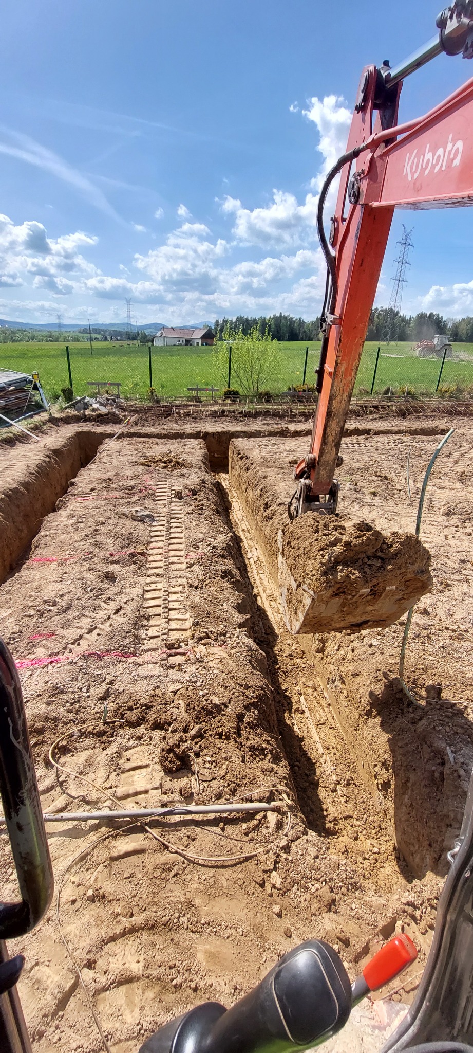 Pomarańczowa koparka Kubota w trakcie wykopu wąskiego rowu w ziemi na tle zielonego pola i błękitnego nieba z chmurami. Widoczne ślady gąsienic.