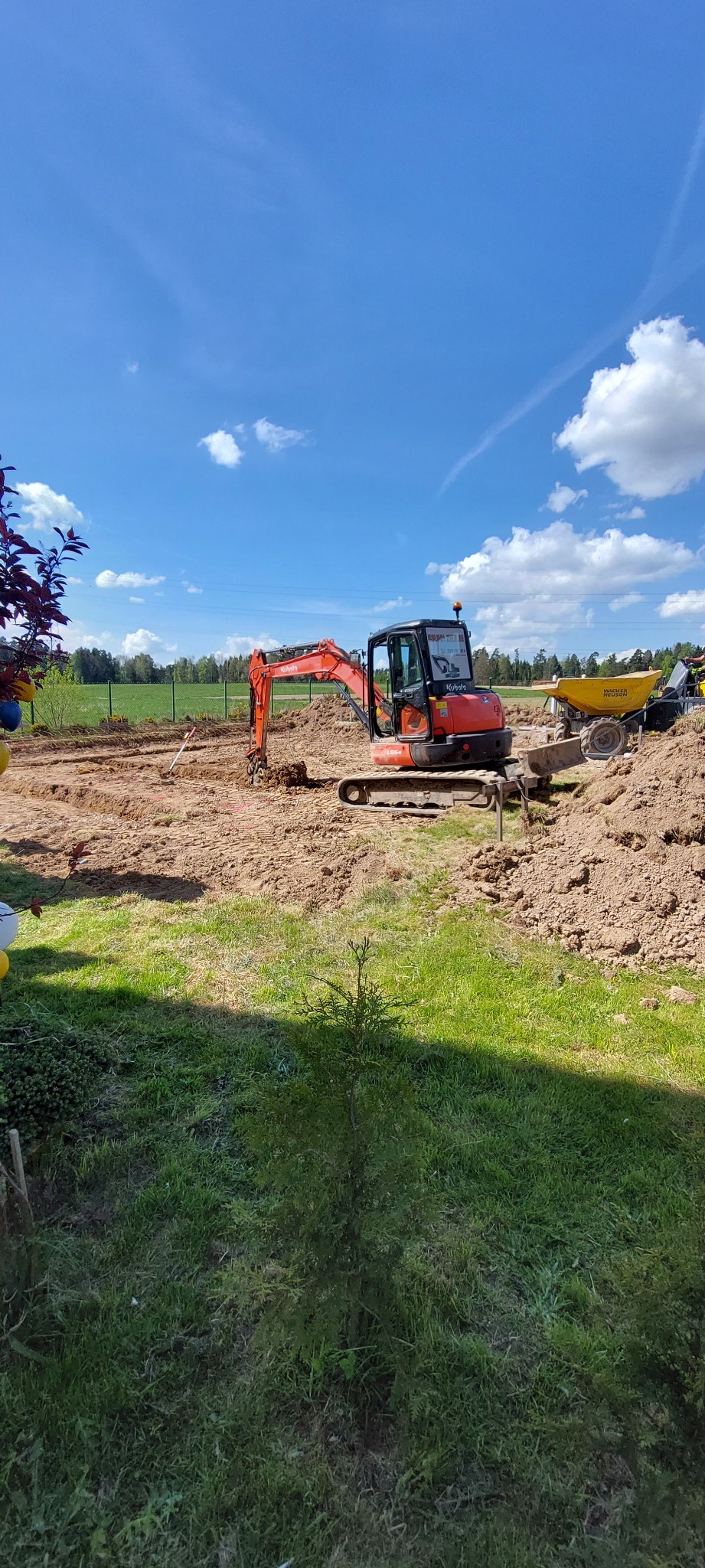 Pomarańczowa mini koparka Kubota i wywrotka Wacker Neuson na placu budowy, wykop pod fundamenty, błękitne niebo z chmurami w tle.