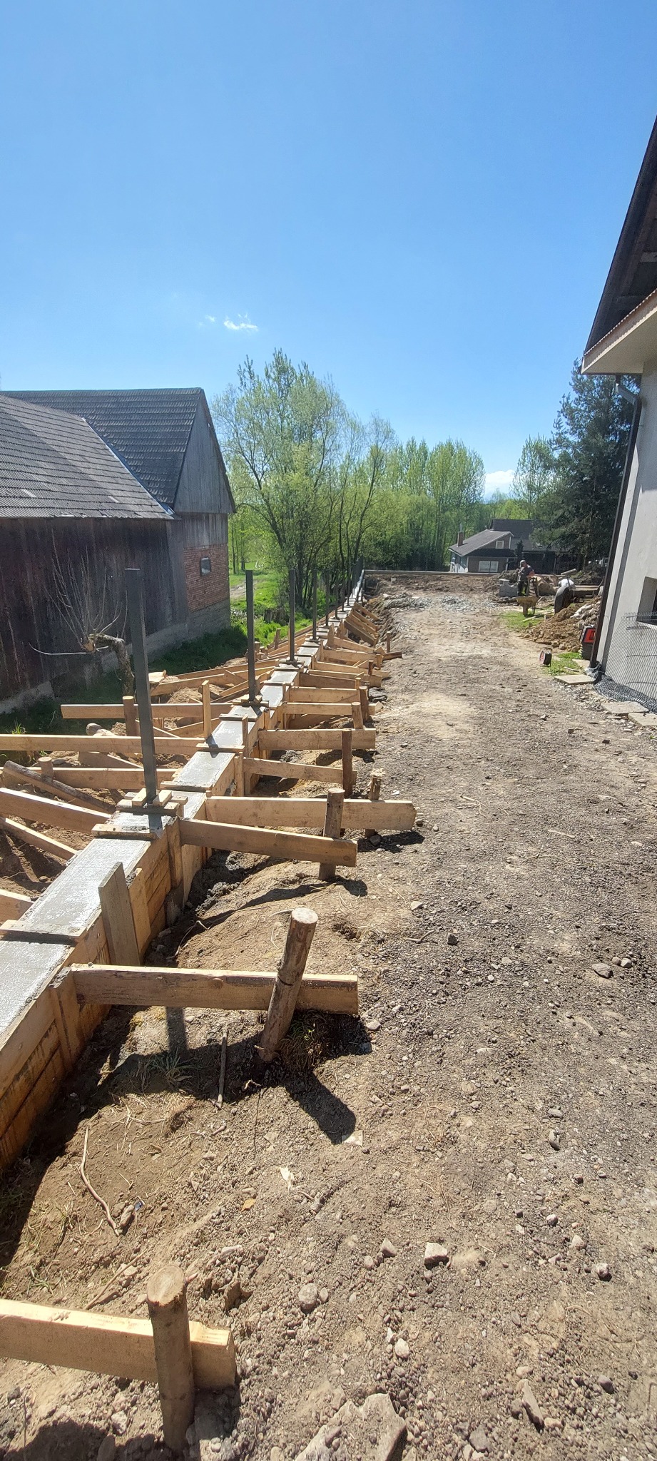 Fundamenty w drewnianym szalunku, częściowo zalane betonem, widoczne stalowe pręty. W tle budynki i drzewa, słoneczny dzień.