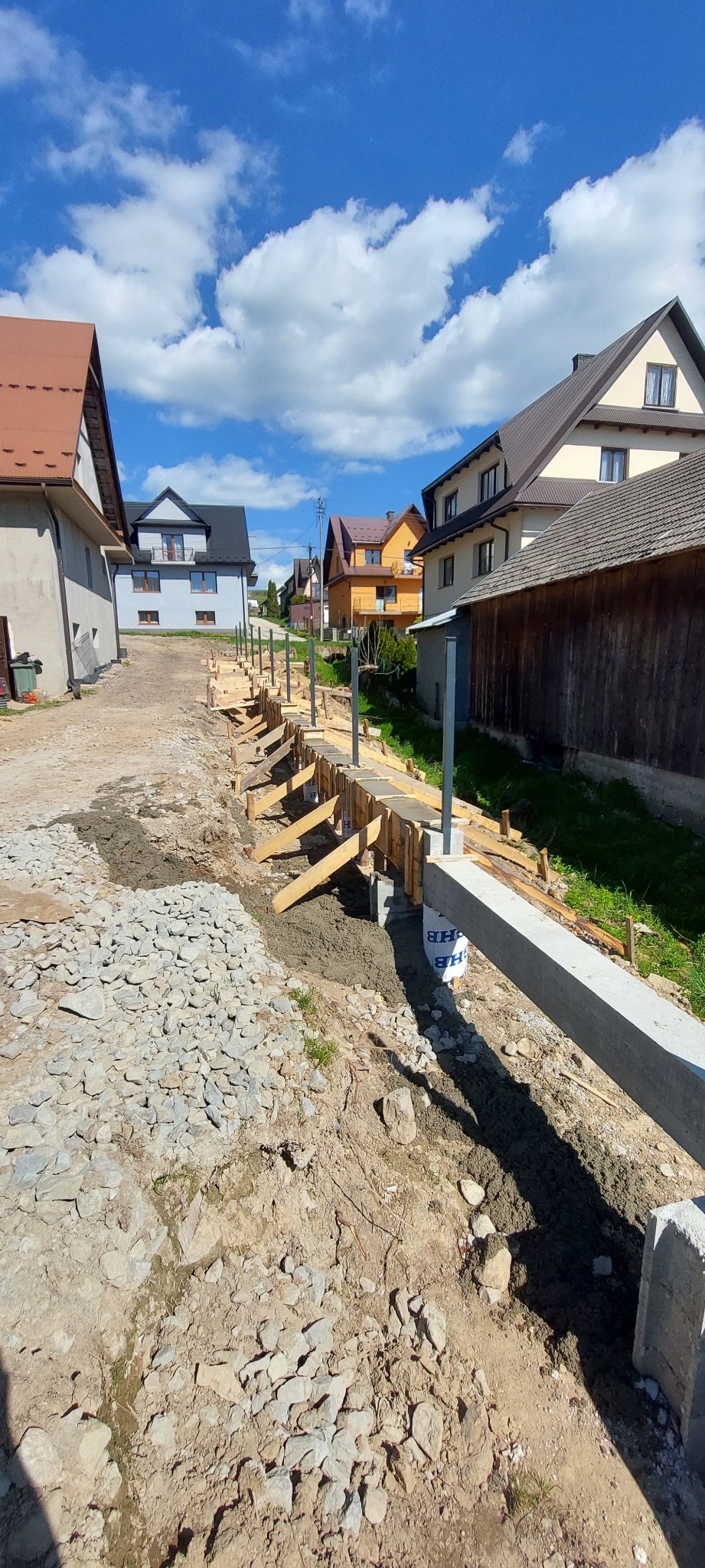 Fundamenty pod ogrodzenie w trakcie budowy, szalunek z drewnianych desek, stalowe słupki, kamienie, domy w tle, błękitne niebo z chmurami.