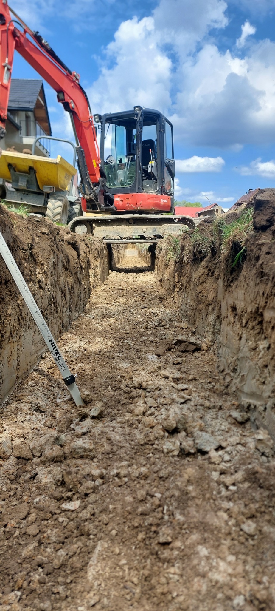 Wąski wykop w ziemi z widocznym czerwonym minikoparką i żółtą wywrotką w tle, z białą łatą niwelacyjną w lewym dolnym rogu. Błękitne niebo z chmurami w tle.