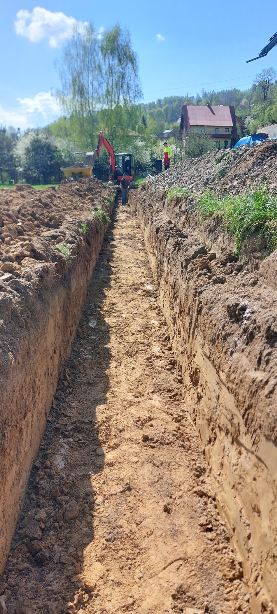 Długi wykop w ziemi, w tle koparka i pracownicy. Wykop świeży, z wyraźnymi śladami pracy maszyny. Praca w toku, teren zielony w tle.