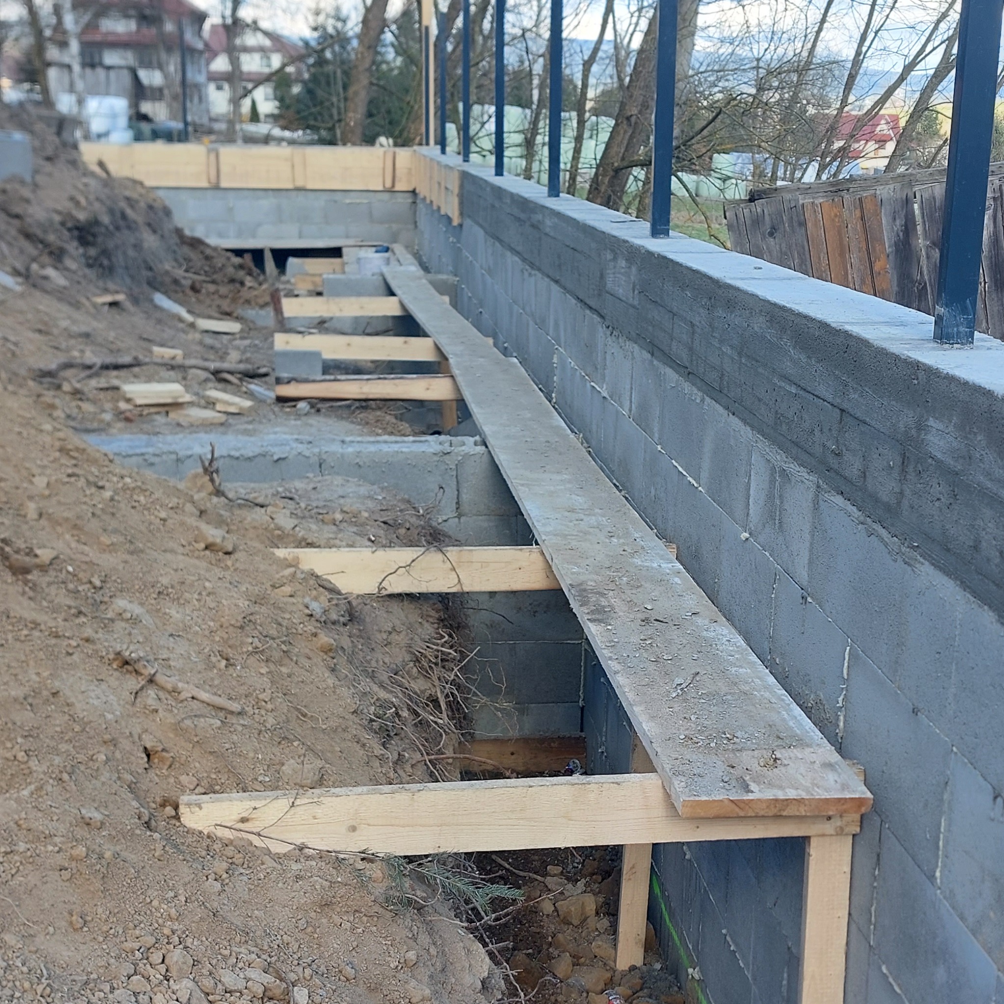 Fundament z bloczków betonowych, częściowo zasypany ziemią, z drewnianym szalunkiem. Widoczne zbrojenie pionowe. Tło stanowią drzewa i zabudowania.
