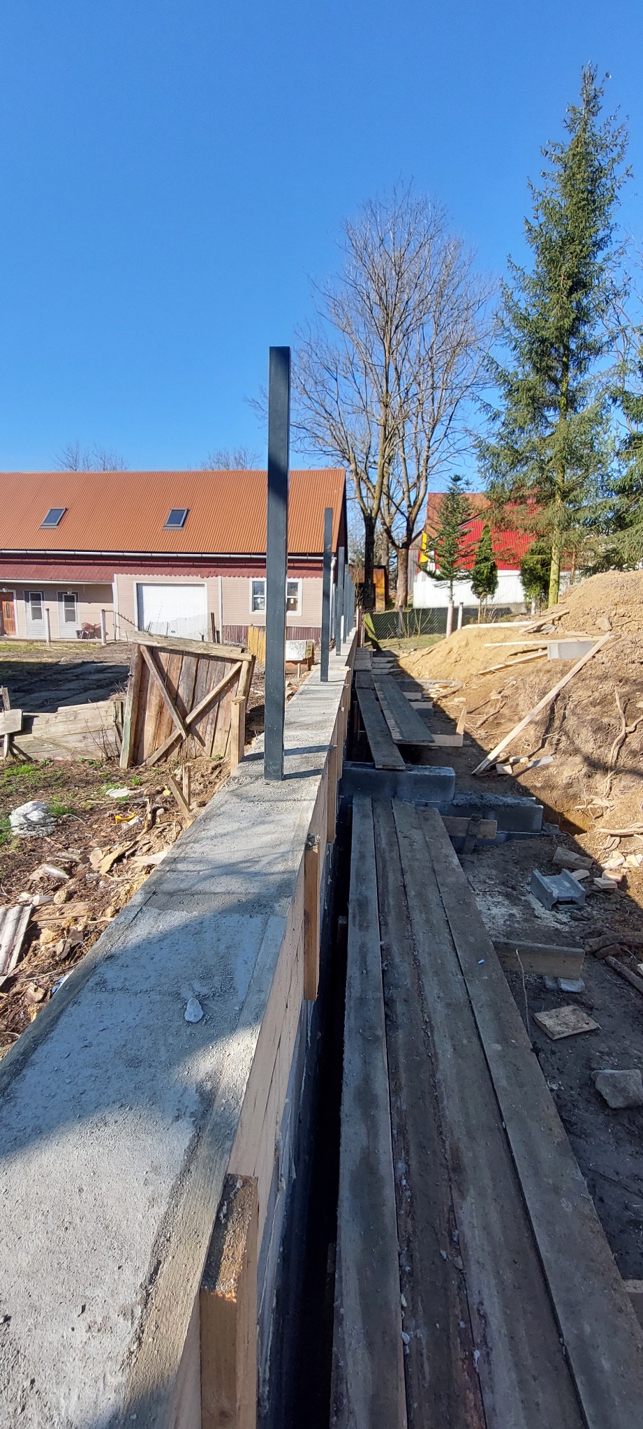 Wąski, betonowy fundament z wystającymi, czarnymi słupkami, drewnianym szalunkiem i ziemią w tle, w słoneczny dzień. Widok z perspektywy.