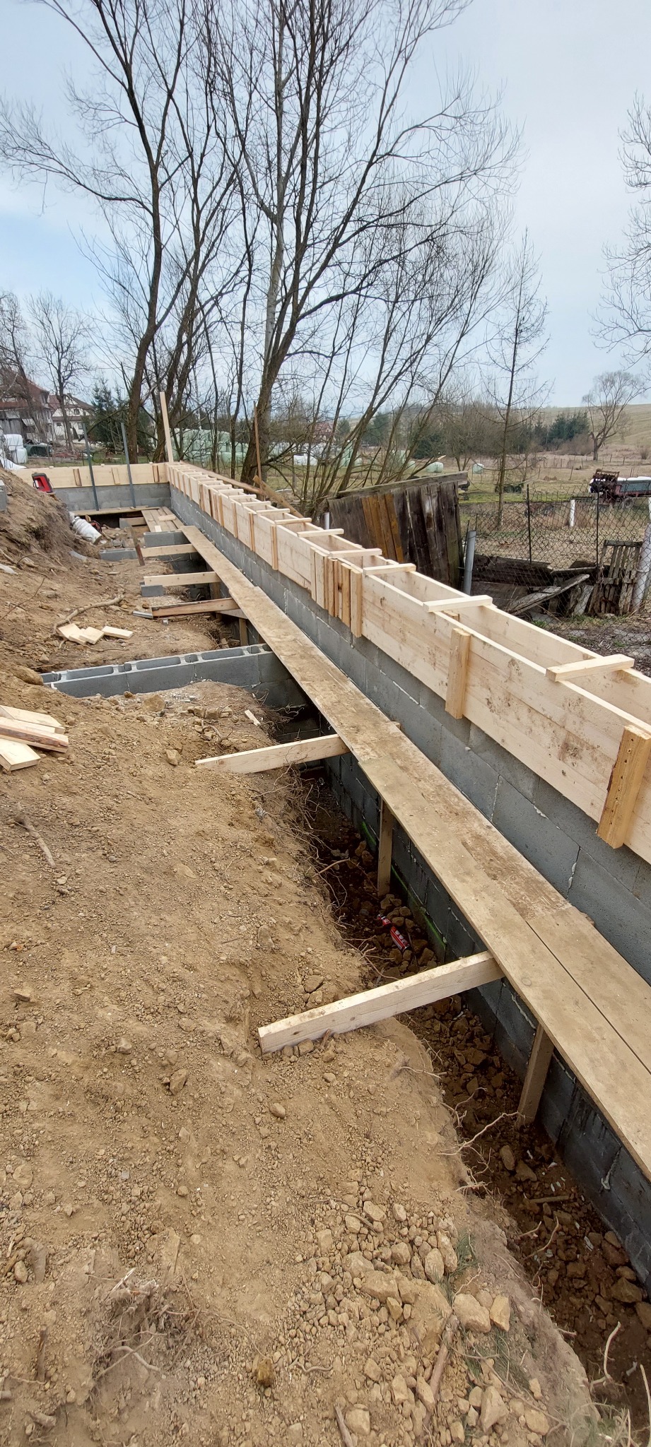 Szare bloczki fundamentowe z drewnianym szalunkiem na budowie domu jednorodzinnego, widok z góry na wykop z ziemią i drzewami w tle.