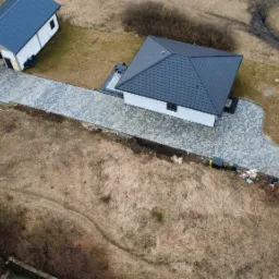 Widok z drona na posesję z ułożoną kostką brukową: podjazd, taras przy domu i ścieżka do furtki. Kostka w odcieniach szarości. Budynki z ciemnymi dachami. Okolica w stanie surowym.