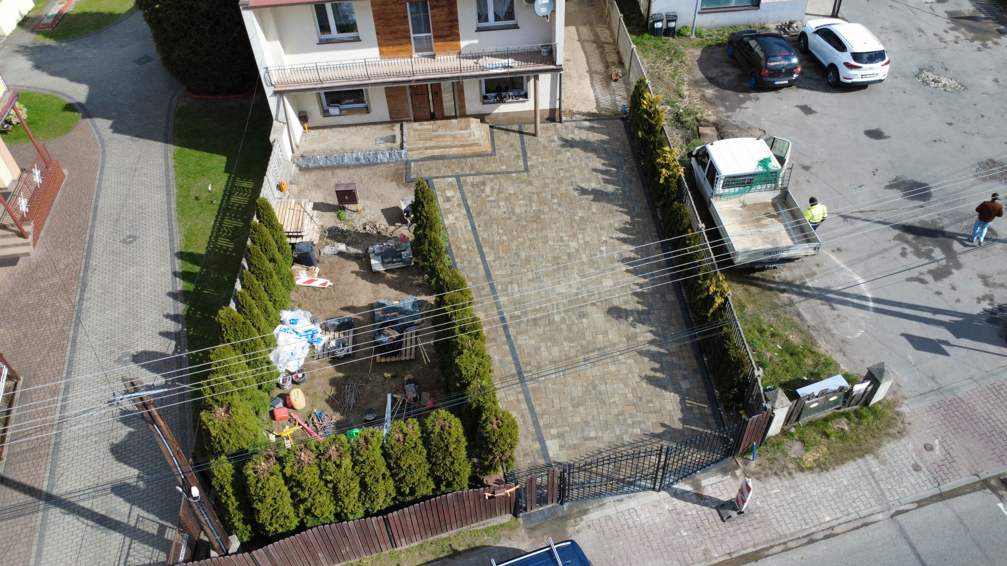 Widok z góry na posesję z nową, ułożoną kostką brukową na podjeździe, obramowaną tujami i ogrodzeniem. Część terenu jeszcze w trakcie prac, widoczne materiały i pracownicy.