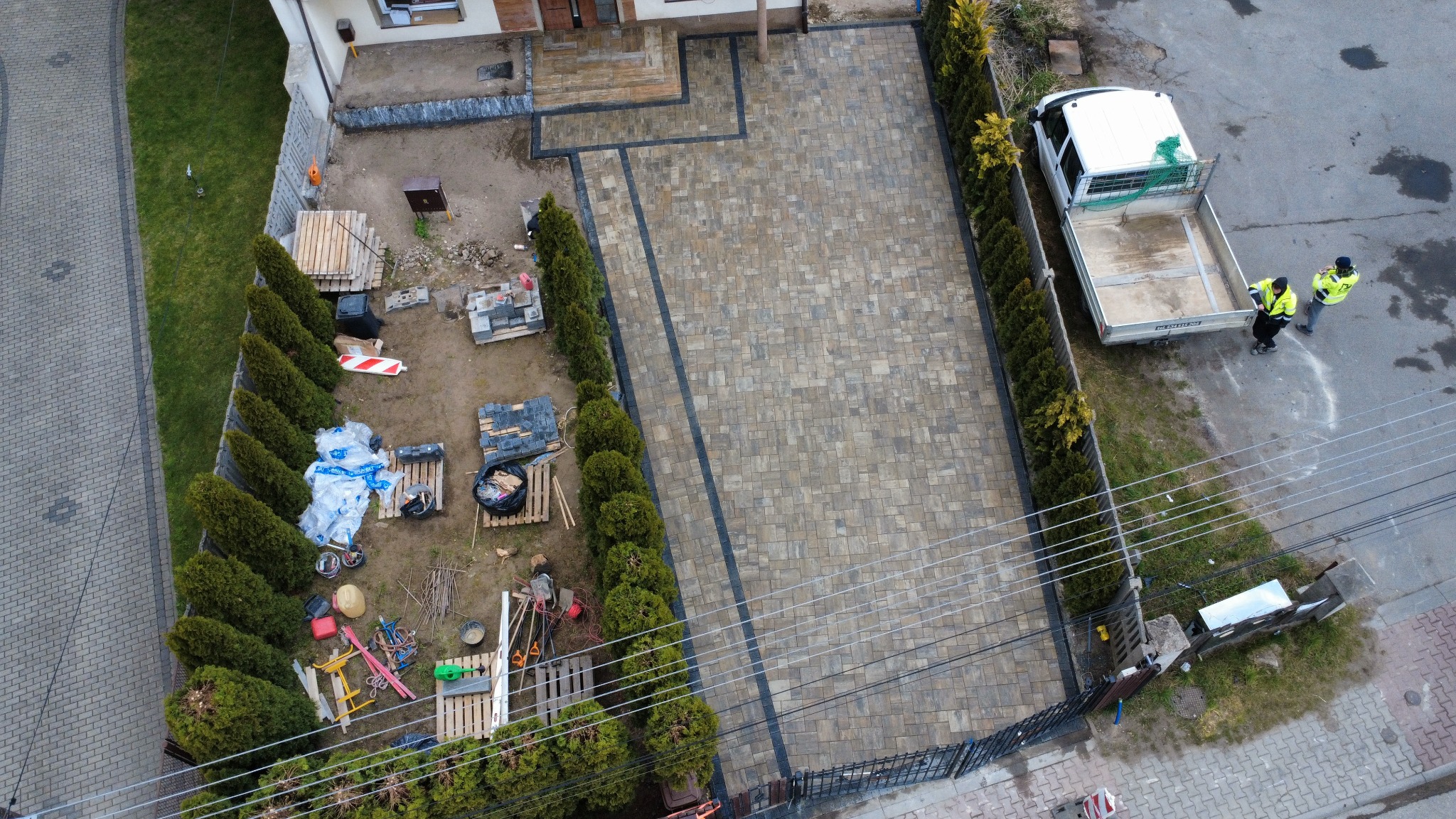 Widok z góry na nową, beżowo-szarą kostkę brukową z ciemnymi obrzeżami, otoczoną tujami i ogrodzeniem. W tle zaparkowany samochód dostawczy i materiały budowlane.