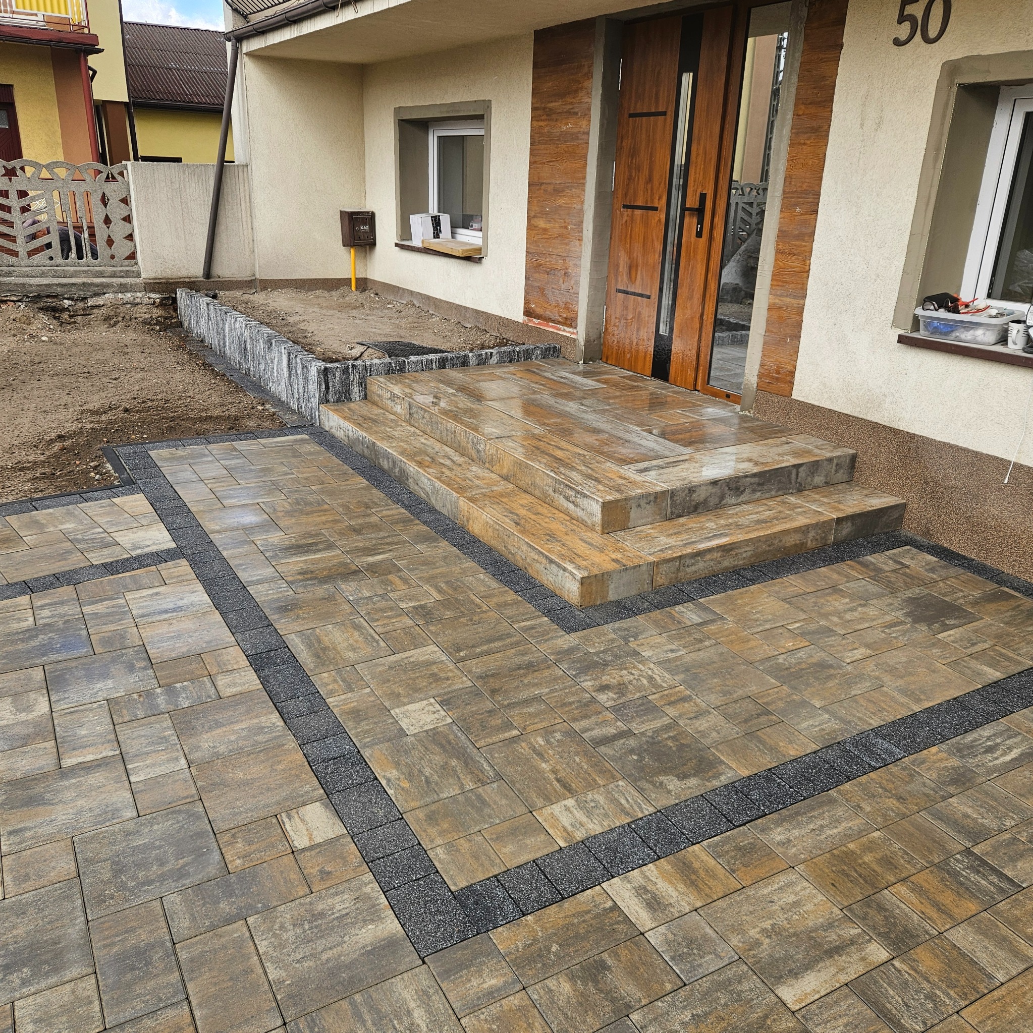Ułożona kostka brukowa w odcieniach brązu i szarości, tworząca ścieżkę i schody przed domem z numerem 50, z widoczną ziemią w tle.