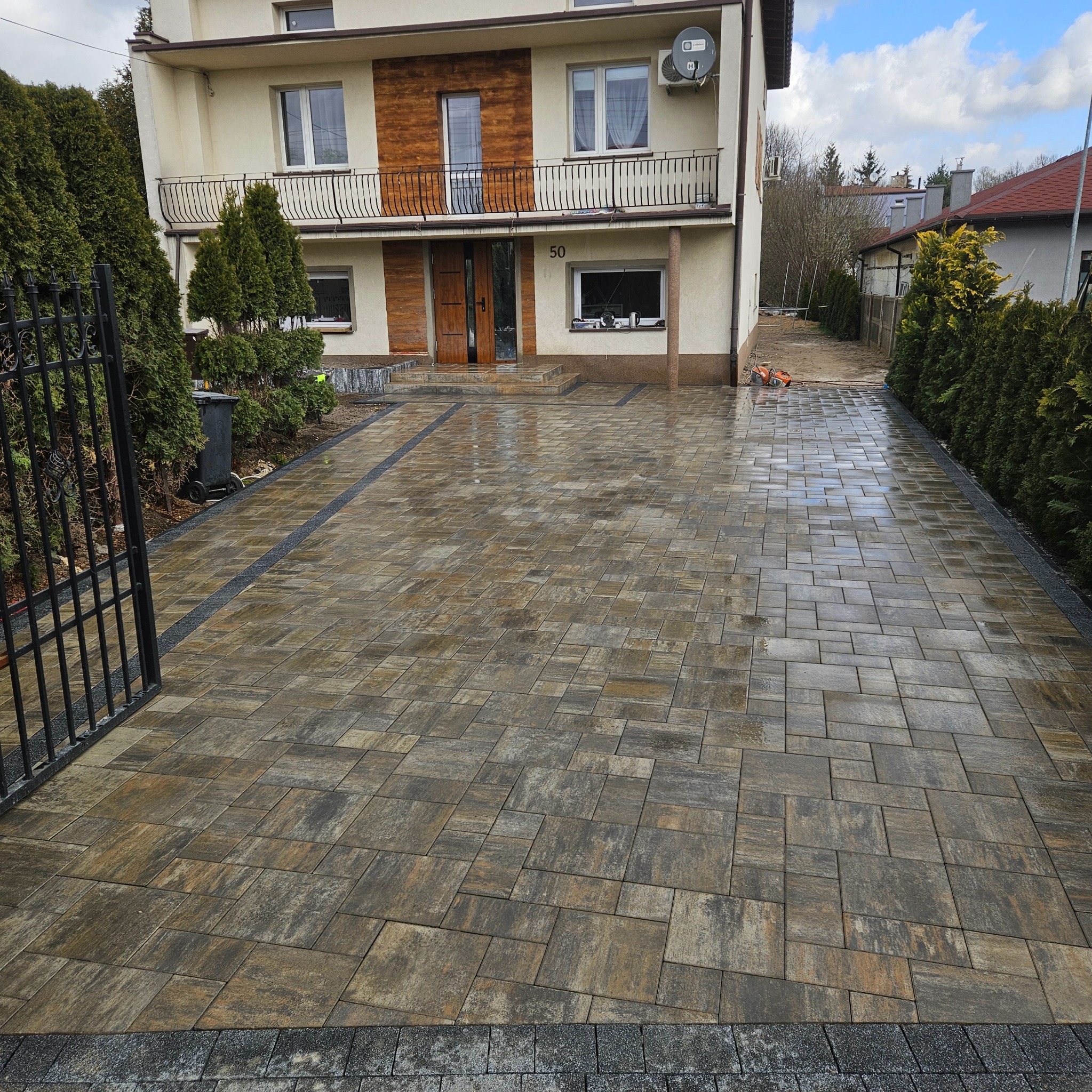 Nowa, brązowo-szara kostka brukowa na podjeździe domu z numerem 50, obrzeża z szarej kostki. Widoczny fragment bramy i maszyna do cięcia kostki w tle.