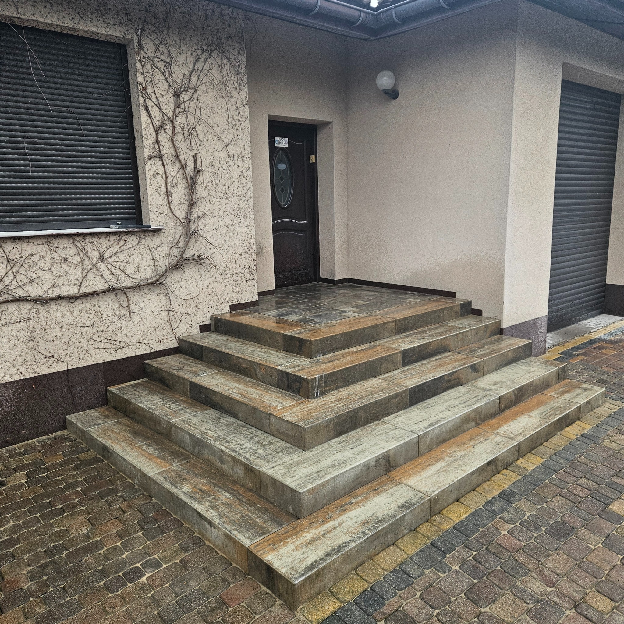 Betonowe schody wejściowe z kostki brukowej w odcieniach szarości i brązu, prowadzące do drzwi wejściowych budynku z elewacją w jasnym odcieniu beżu.