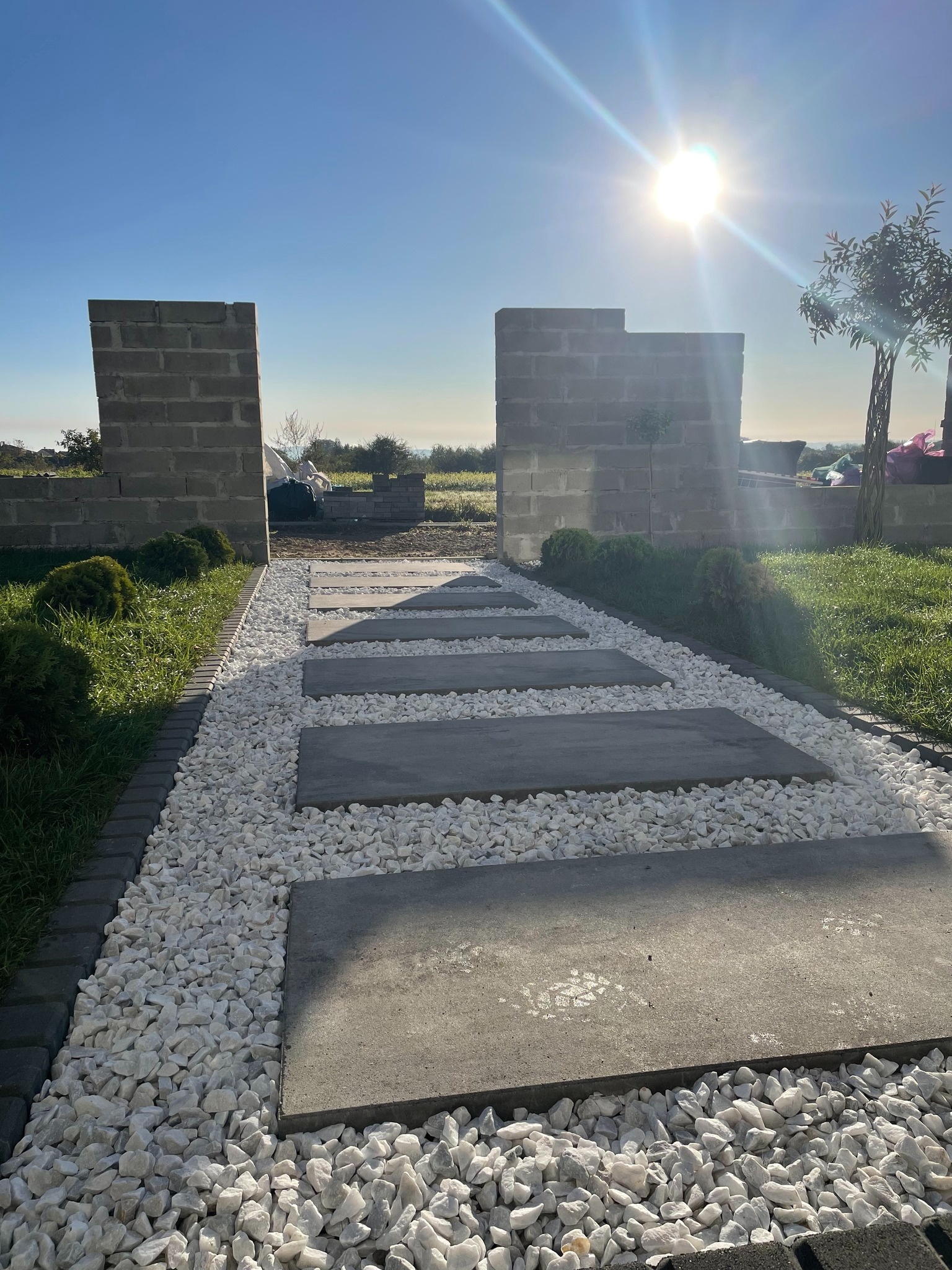 Ścieżka z betonowych płyt i białego grysu, obramowana kostką, prowadząca przez ogród z niskimi murkami. Słoneczny dzień.