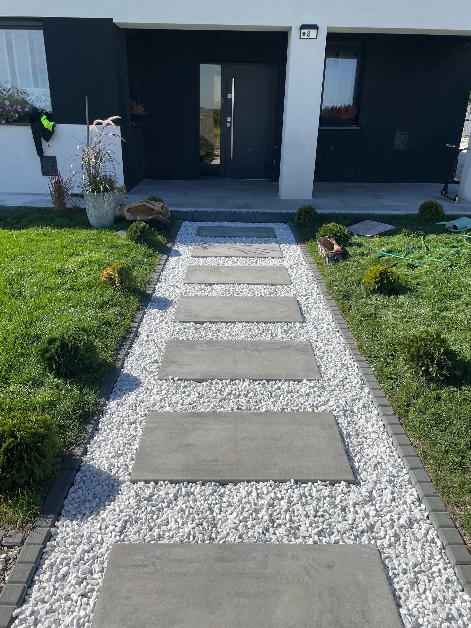 Nowoczesna ścieżka z płyt betonowych i białego grysu prowadząca do domu z czarno-białą elewacją. Dekoracyjne krzewy i trawnik po bokach.