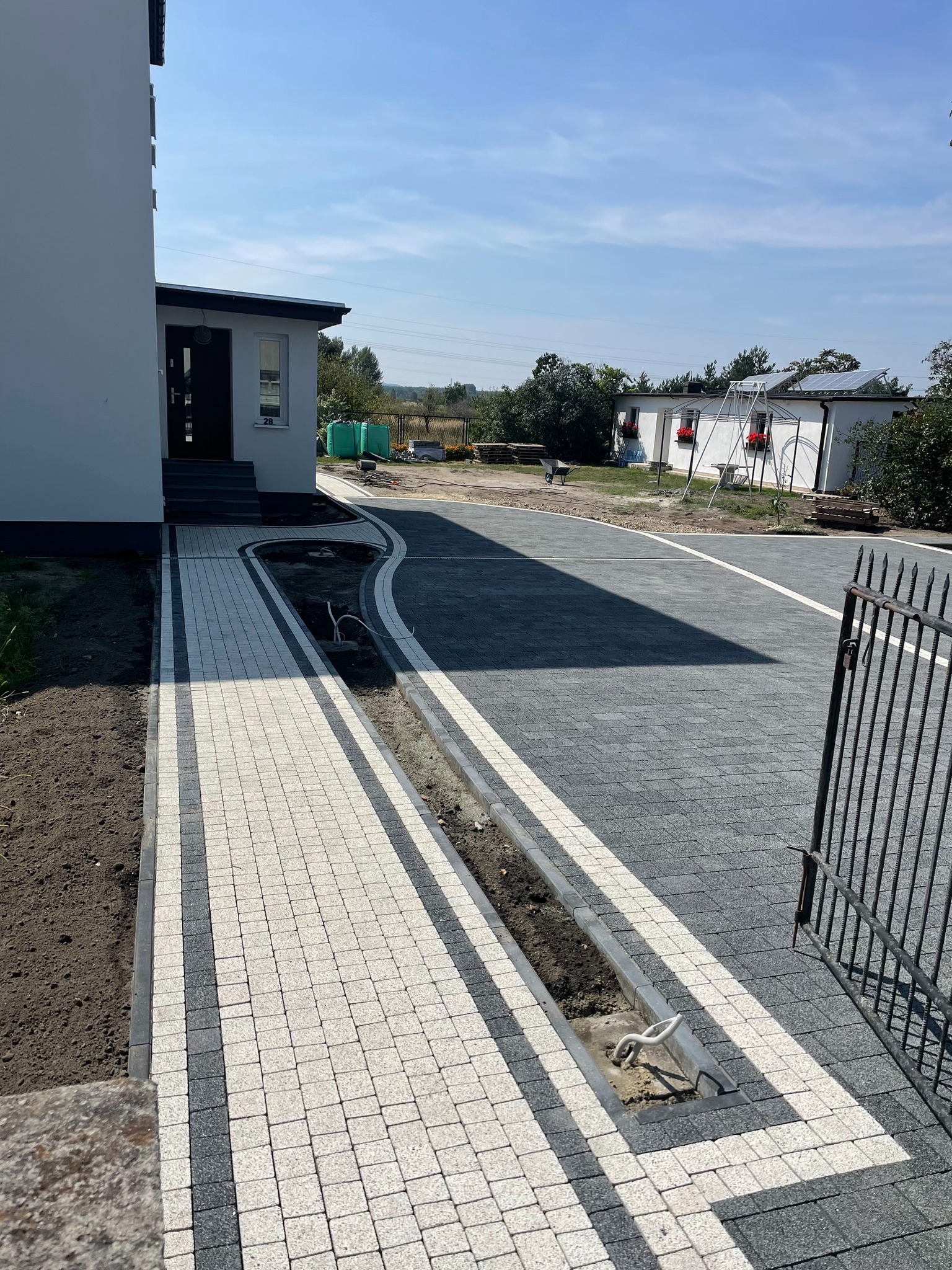 Ułożona kostka brukowa w dwóch odcieniach szarości, tworząca wzór chodnika prowadzącego do domu. Widoczne korytka odwadniające i fragment metalowej bramy.
