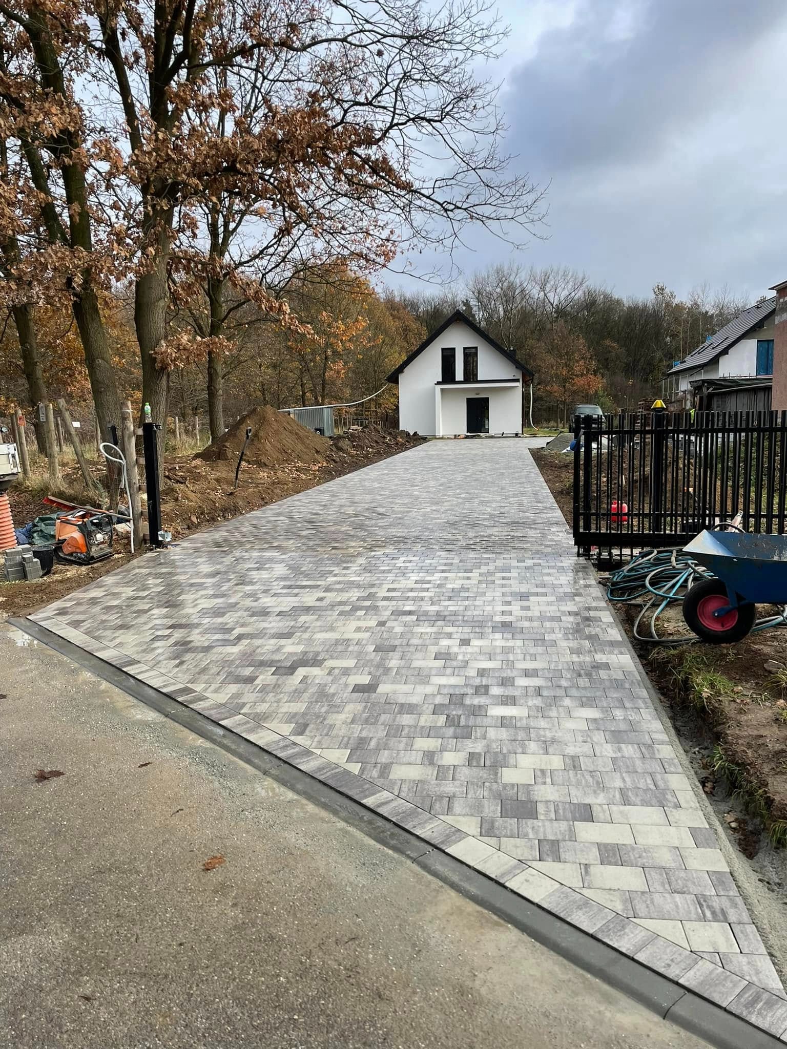 Ułożona, wzorzysta kostka brukowa prowadząca do białego domu z czarnym dachem. Widoczne narzędzia i maszyna budowlana po lewej stronie. Jesienne drzewa w tle.