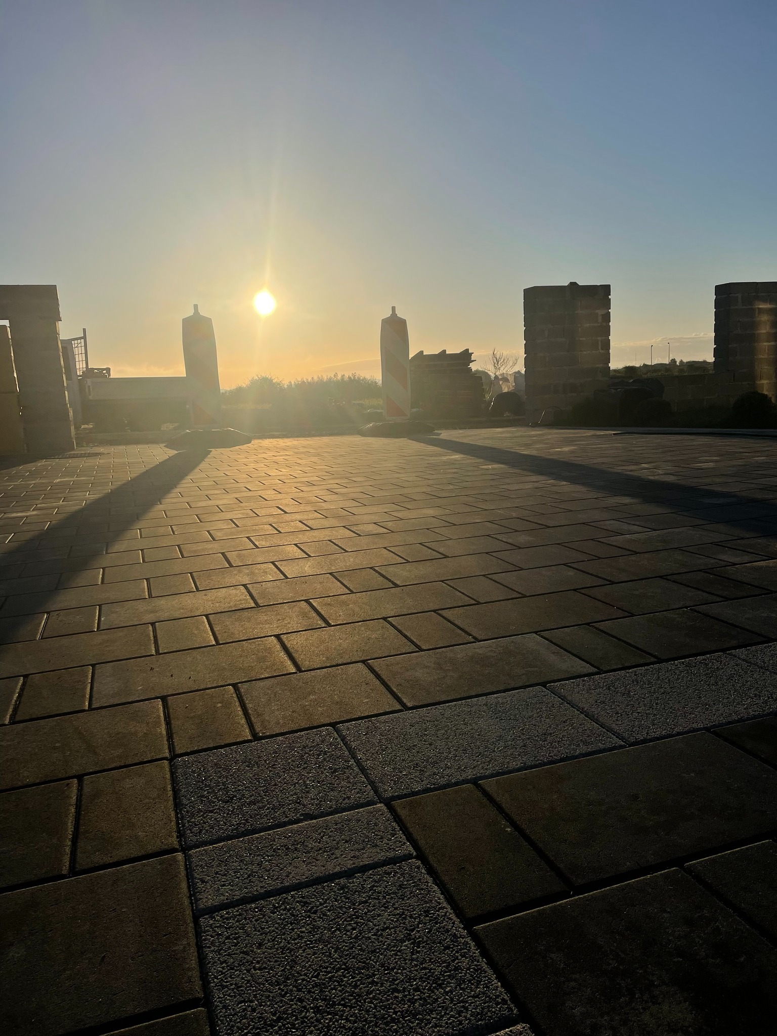 Ułożona kostka brukowa w ciepłym świetle wschodzącego słońca, z widocznymi elementami roboczymi w tle i paletami kostki po bokach.