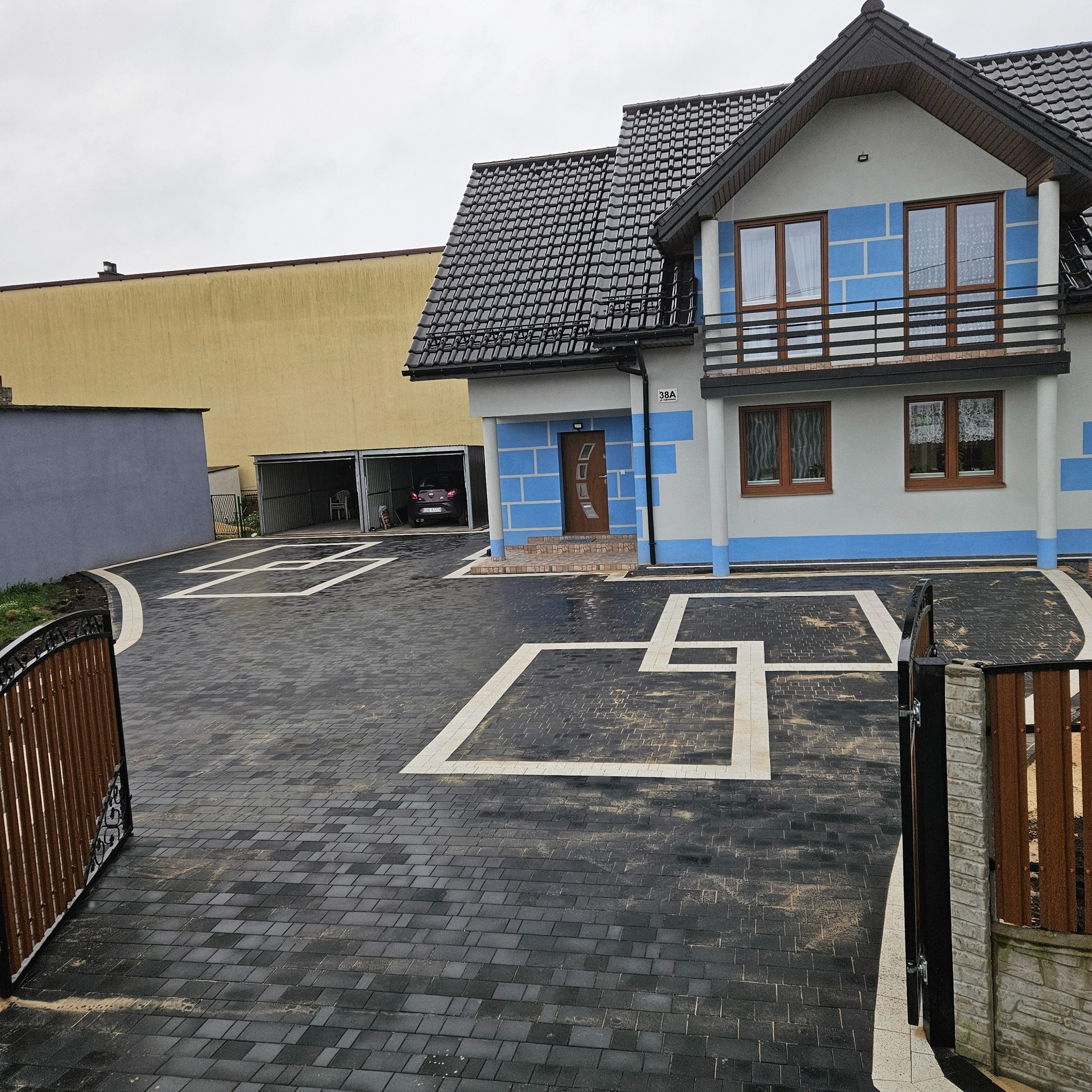 Nowoczesny podjazd z ciemnej kostki brukowej z białymi akcentami, prowadzący do domu z balkonem. Widoczna brama wjazdowa i garaż.