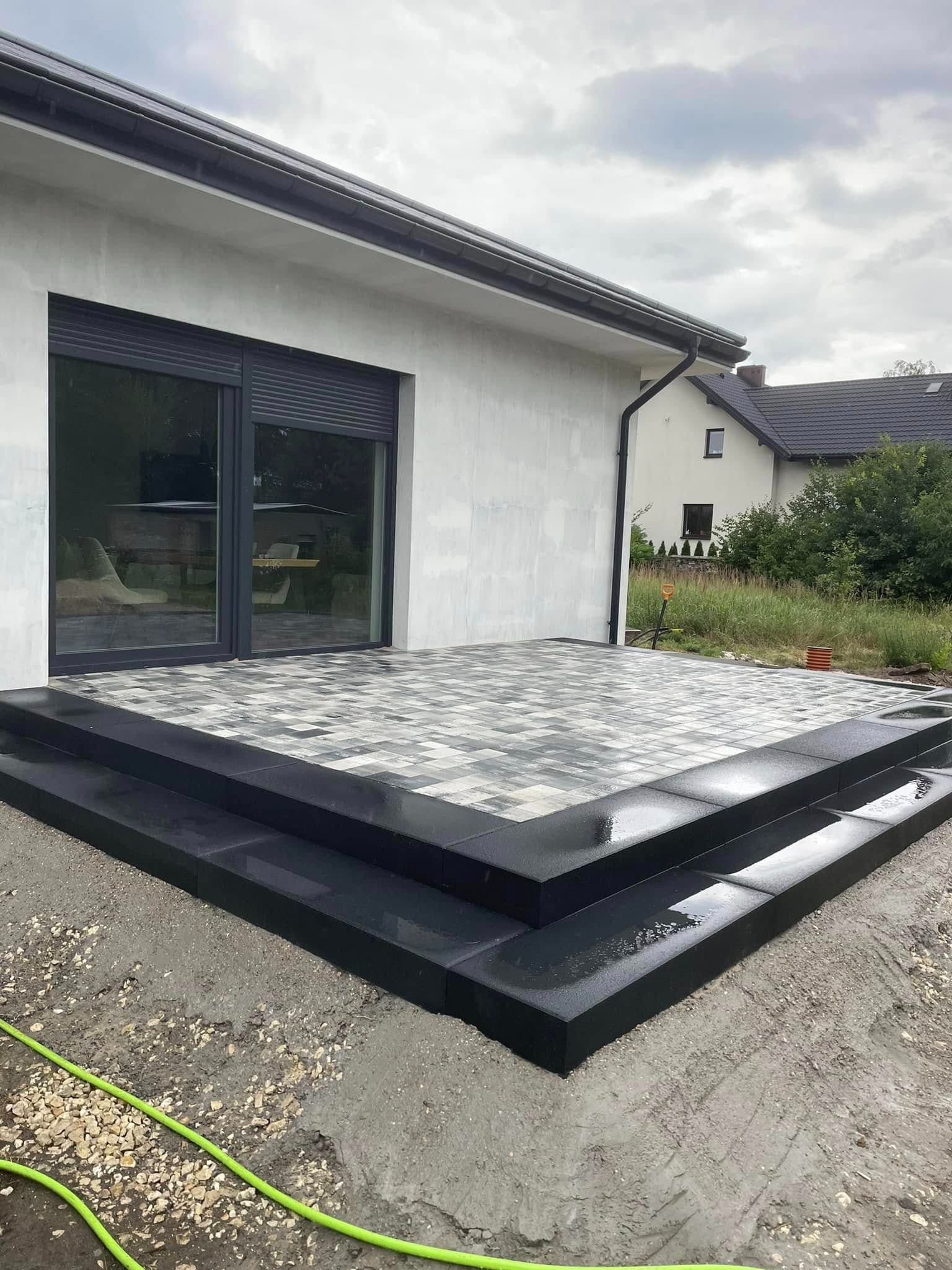 Nowoczesny taras z kostki brukowej w Psarach, z niskimi schodami. Szaro-biała kostka ułożona wzorem, ciemne obrzeża. Widok na dom i ogród w tle.