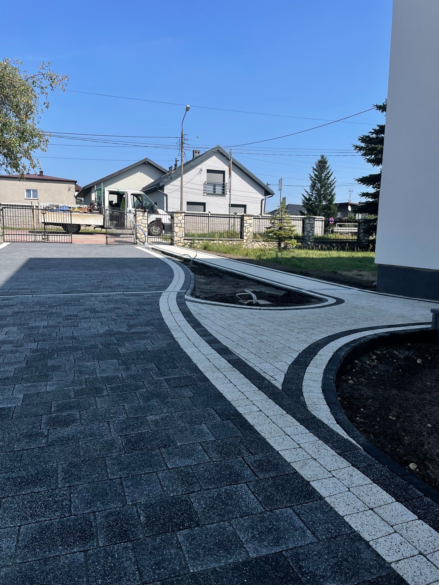 Nowo ułożona, dwukolorowa kostka brukowa przed domem z podjazdem i rabatami. Widoczny fragment budynku i samochód dostawczy w tle. Słoneczny dzień.