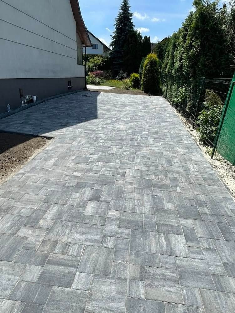 Ułożona szara kostka brukowa na podjeździe domu jednorodzinnego, widok z perspektywy. Kostka ułożona w nieregularny wzór, obrzeża z ziemi i trawy.