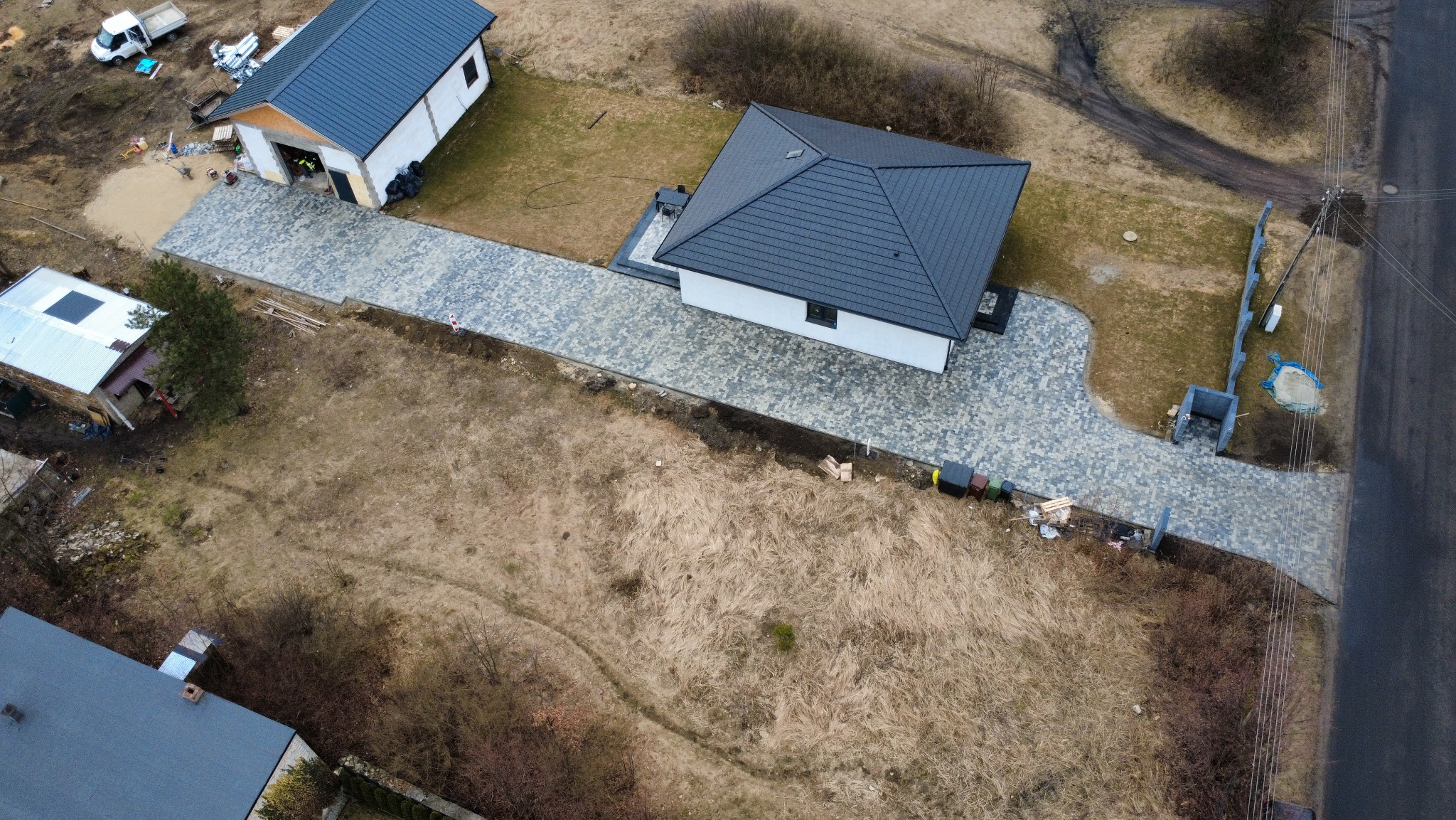 Widok z drona na posesję z ułożoną kostką brukową: podjazd, taras przy domu i ścieżka do furtki. Kostka w odcieniach szarości. Budynki z ciemnymi dachami. Okolica w stanie surowym.