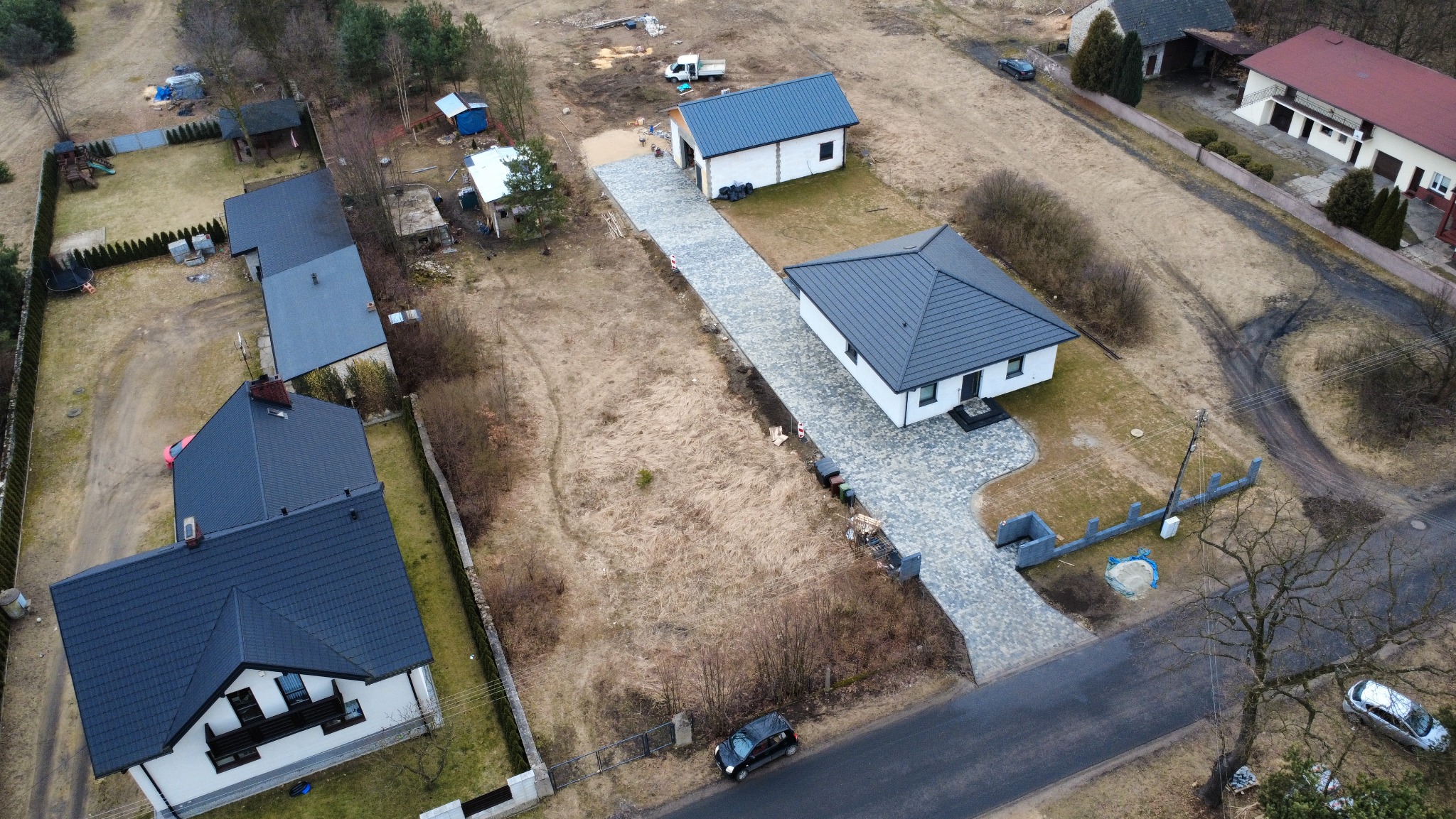 Widok z drona na posesję z nowo ułożoną, szarą kostką brukową prowadzącą do domu i garażu; otoczenie w stanie surowym, w tle zabudowa jednorodzinna.