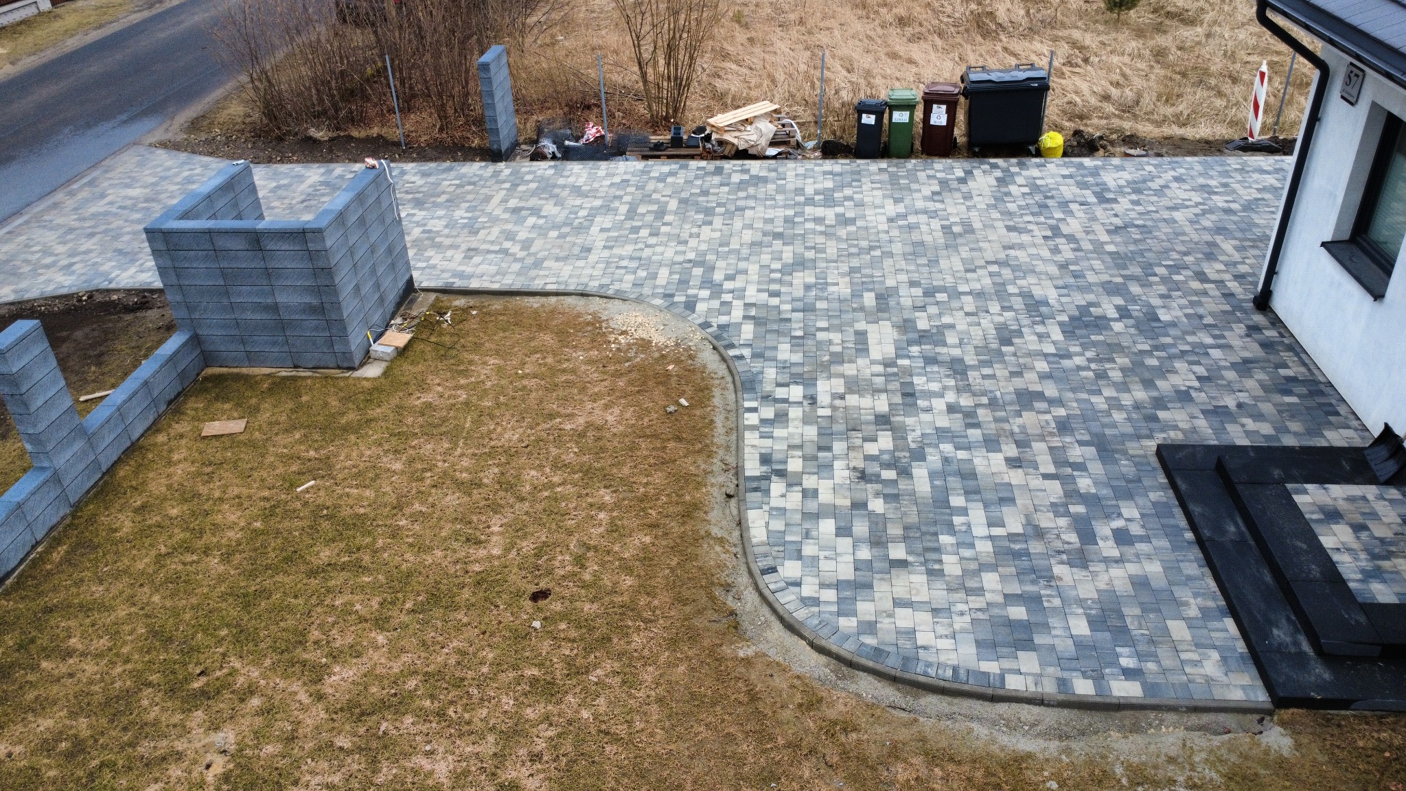 Ułożona kostka brukowa w odcieniach szarości tworząca podjazd i ścieżkę, widok z góry. Fragment murku z bloczków betonowych obok trawnika.