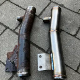 Porównanie: skorodowana rura metalowa obok nowej, spawanej rury z uchwytem, leżących na szarym bruku.