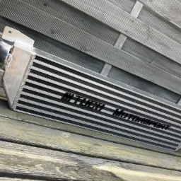 Aluminiowy intercooler z czarnym napisem 'HT-TECH Motorsport' leżący na szarych, drewnianych deskach na zewnątrz.