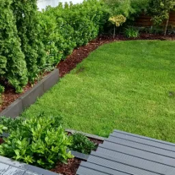 Widok z góry na świeżo założony ogród z idealnie przystrzyżonym trawnikiem, obrzeżami z kory i niskimi krzewami, widziany z drewnianego tarasu.