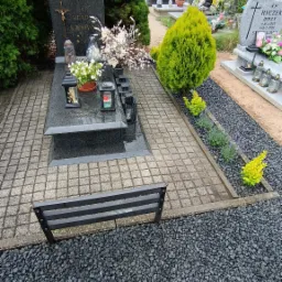 Aranżacja grobu z czarnego granitu, otoczona szarą kostką brukową i rabatą z czarnych kamieni, na której rosną karłowate iglaki i lawenda. W tle widoczne inne nagrobki i drzewa.