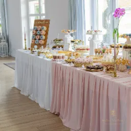 Bogato zastawiony stół ze słodkościami: donuty na drewnianym stojaku z napisem, ciasta, desery w szklankach, ciasteczka, torty, złote patery, świeczniki, różowe kwiaty, biało-różowy obrus z logo...