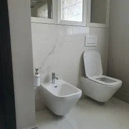 Minimalistyczna łazienka z białymi płytkami imitującymi marmur, wiszącą toaletą i bidetem oraz chromowaną armaturą, uzupełniona o dozownik mydła i lustrzane szafki.