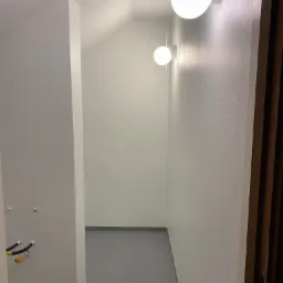 Wnętrze korytarza z szarą podłogą, białymi ścianami o delikatnej strukturze i dwoma okrągłymi lampami ściennymi emitującymi ciepłe światło, widoczne wystające rury instalacji.